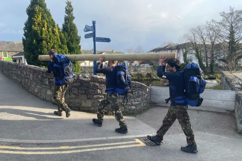 Three people carrying a large log over a bridge in Keswick