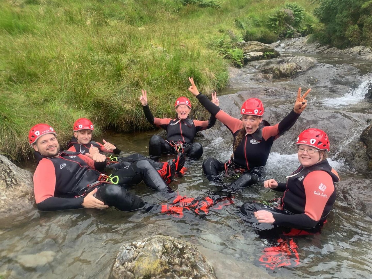 Family of 5 people in a shallow river, showing thumbs up and peace signs